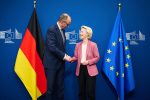 Friedrich Merz and Ursula von der Leyen shaking hands between the German and EU flags at the European Commission in Brussels